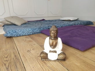 A serene room with a wooden floor, featuring a small statue of a Buddha sitting in a meditative pose. Behind the statue, there are cushions and a blue patterned mat arranged on the floor, creating a peaceful and relaxing atmosphere.