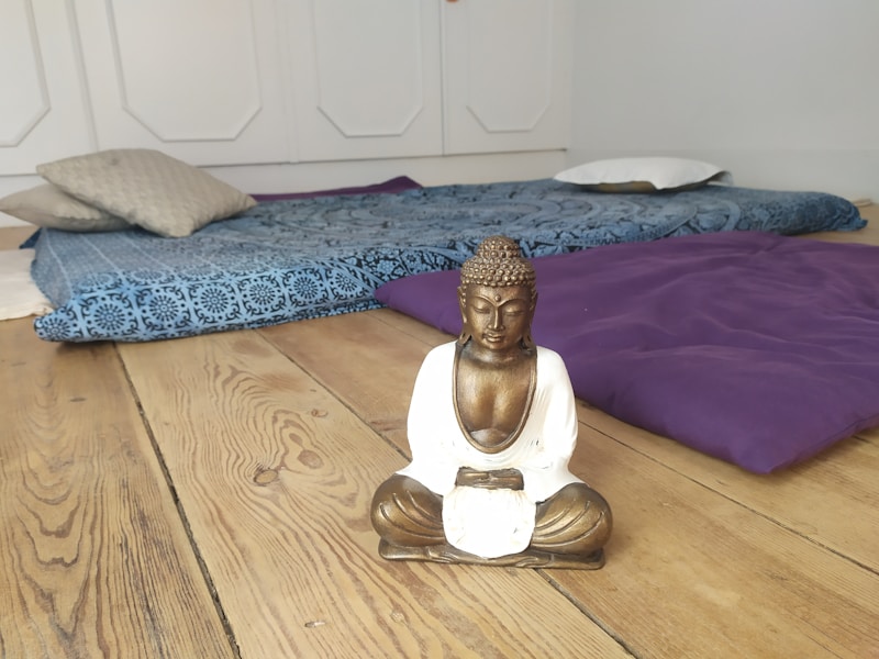 A serene room with a wooden floor, featuring a small statue of a Buddha sitting in a meditative pose. Behind the statue, there are cushions and a blue patterned mat arranged on the floor, creating a peaceful and relaxing atmosphere.
