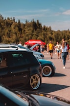 Several cars are parked in an outdoor area, with a group of people walking and interacting in the background. The cars are prominently displayed, and some have custom modifications, like lowered suspension and aftermarket wheels. A red tent or sign with indistinguishable text is visible in the background, set against a backdrop of trees and a clear sky.