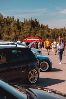 Several cars are parked in an outdoor area, with a group of people walking and interacting in the background. The cars are prominently displayed, and some have custom modifications, like lowered suspension and aftermarket wheels. A red tent or sign with indistinguishable text is visible in the background, set against a backdrop of trees and a clear sky.