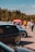 Several cars are parked in an outdoor area, with a group of people walking and interacting in the background. The cars are prominently displayed, and some have custom modifications, like lowered suspension and aftermarket wheels. A red tent or sign with indistinguishable text is visible in the background, set against a backdrop of trees and a clear sky.