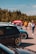 Several cars are parked in an outdoor area, with a group of people walking and interacting in the background. The cars are prominently displayed, and some have custom modifications, like lowered suspension and aftermarket wheels. A red tent or sign with indistinguishable text is visible in the background, set against a backdrop of trees and a clear sky.