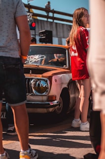 A group of people, some partly visible, are gathered around a vintage car with rust patches and a customized headlight. One person in the foreground wears a red sports jersey, while another person is seen from behind wearing shorts. The environment appears to be a casual outdoor event or car show.