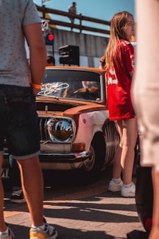 A group of people, some partly visible, are gathered around a vintage car with rust patches and a customized headlight. One person in the foreground wears a red sports jersey, while another person is seen from behind wearing shorts. The environment appears to be a casual outdoor event or car show.