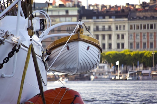 A lifeboat is suspended from a ship, with an urban waterfront and buildings in the background. The image captures nautical equipment and architectural structures under a clear sky.