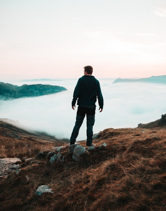 man in black jacket standing on brown rock near body of water during daytime in Lake District National Park United Kingdom