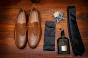 A pair of tan leather loafers with tassels are placed on a wooden surface. Alongside them is a black tie, a matching pocket square, a small bottle of dark-colored cologne, and a boutonniere with white and purple flowers and green leaves.
