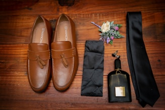 A pair of tan leather loafers with tassels are placed on a wooden surface. Alongside them is a black tie, a matching pocket square, a small bottle of dark-colored cologne, and a boutonniere with white and purple flowers and green leaves.