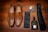 A pair of tan leather loafers with tassels are placed on a wooden surface. Alongside them is a black tie, a matching pocket square, a small bottle of dark-colored cologne, and a boutonniere with white and purple flowers and green leaves.
