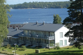 A modern two-story building with large windows and a dark gray roof is surrounded by lush greenery and overlooks a serene body of water. The shoreline is lined with dense trees, and the vibrant landscape includes a well-maintained lawn and a few smaller structures with red roofs nearby.
