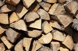 A pile of neatly stacked firewood with visible cut surfaces displaying rings and a variety of brown and beige tones. The logs are arranged in a compact manner, with rough, textured surfaces and some dusting of bark still attached.