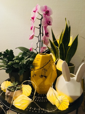 A vibrant arrangement featuring a pink orchid with multiple blooms in the center, surrounded by various green potted plants. A prominent yellow vase decorated with cartoon characters sits beside a white vase shaped like a rabbit. Illuminating the scene are string lights with yellow geometric paper shades, arranged aesthetically around the objects.