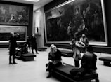 A group of people is inside an art gallery, observing large paintings on the walls. Some individuals are sitting on benches, while others walk around. A man is taking a photo, and another person appears to be reading a booklet. The gallery features dramatic artwork, and the space is well-lit with modern design elements.