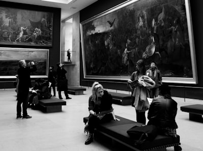 A group of people is inside an art gallery, observing large paintings on the walls. Some individuals are sitting on benches, while others walk around. A man is taking a photo, and another person appears to be reading a booklet. The gallery features dramatic artwork, and the space is well-lit with modern design elements.