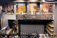 A bar interior featuring a variety of bottled beverages on shelves. Chalkboards display names and prices of different beers. Large images of beers and bar settings adorn the walls. Taps for draft beers are visible at the bottom, and a small shelving unit on the right holds glasses.