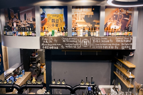 A bar interior featuring a variety of bottled beverages on shelves. Chalkboards display names and prices of different beers. Large images of beers and bar settings adorn the walls. Taps for draft beers are visible at the bottom, and a small shelving unit on the right holds glasses.