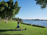 A person is practicing yoga on a mat in a grassy park next to a body of water. The sky is clear and blue, and there are large trees providing shade nearby. A laptop is placed on the grass next to the mat. The scene is peaceful and serene with no visible disturbances.