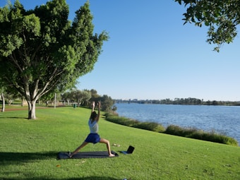 A person is practicing yoga on a mat in a grassy park next to a body of water. The sky is clear and blue, and there are large trees providing shade nearby. A laptop is placed on the grass next to the mat. The scene is peaceful and serene with no visible disturbances.