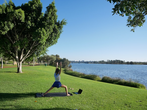 A person is practicing yoga on a mat in a grassy park next to a body of water. The sky is clear and blue, and there are large trees providing shade nearby. A laptop is placed on the grass next to the mat. The scene is peaceful and serene with no visible disturbances.