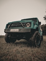 A teal off-road vehicle with large, rugged tires and a prominent front bumper with a license plate reading KL 08 BV 2138 is parked on grass. The sky above is overcast, and a few trees are visible in the background.