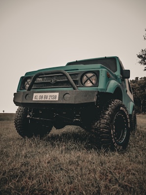 A teal off-road vehicle with large, rugged tires and a prominent front bumper with a license plate reading KL 08 BV 2138 is parked on grass. The sky above is overcast, and a few trees are visible in the background.