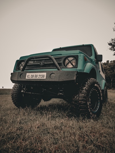 A teal off-road vehicle with large, rugged tires and a prominent front bumper with a license plate reading KL 08 BV 2138 is parked on grass. The sky above is overcast, and a few trees are visible in the background.