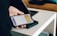 A person is holding a smartphone with a digital payment app open, ready to make a contactless payment on a card reader placed on a white table. The background includes a part of a laptop and a couch.