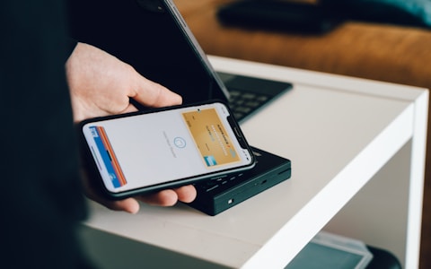 A person is holding a smartphone with a digital payment app open, ready to make a contactless payment on a card reader placed on a white table. The background includes a part of a laptop and a couch.