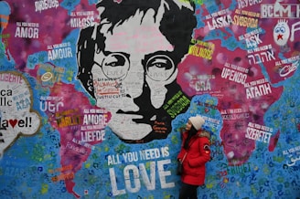 A vibrant graffiti wall featuring a monochromatic portrait surrounded by colorful text. Various expressions of 'All You Need Is Love' appear in multiple languages across the mural, with a woman in a red coat and beanie standing in front, looking up at the artwork.