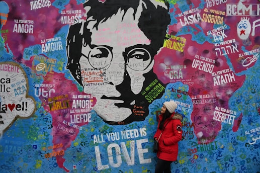 A vibrant graffiti wall featuring a monochromatic portrait surrounded by colorful text. Various expressions of 'All You Need Is Love' appear in multiple languages across the mural, with a woman in a red coat and beanie standing in front, looking up at the artwork.