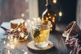 A warm, cozy scene with a glass of tea on a wooden coaster, surrounded by soft, glowing string lights. A dark glass bottle and rustic decor elements, including dried leaves, complement the ambiance.