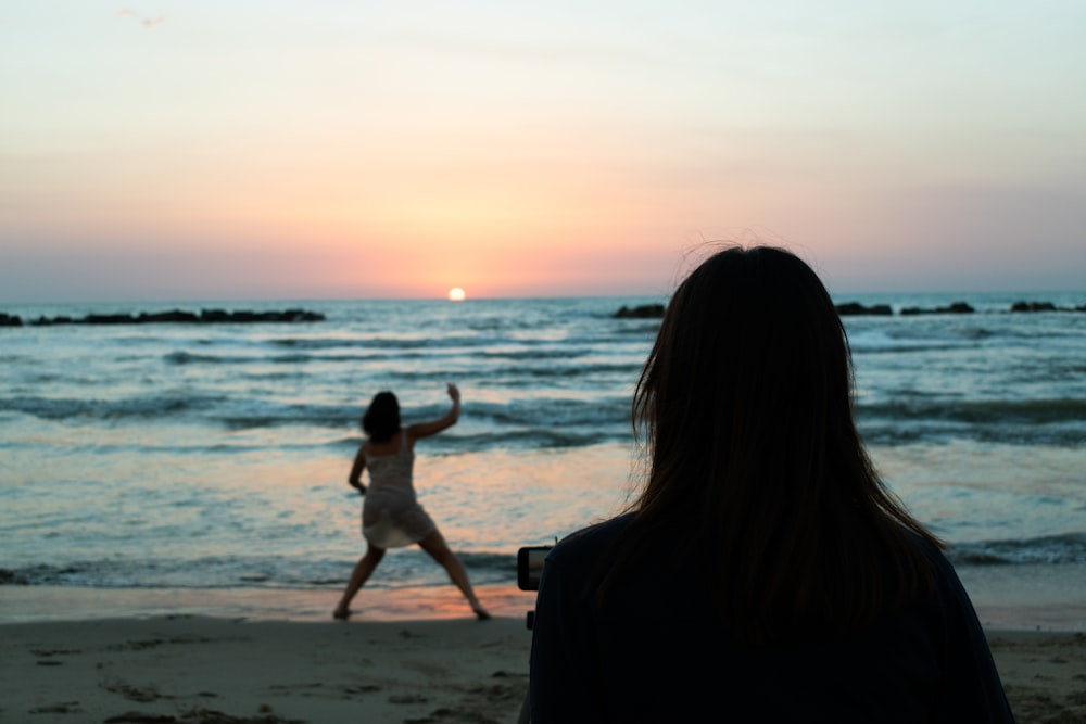 Mujer en vestido negro sosteniendo a una chica en vestido blanco en la playa durante la puesta del sol