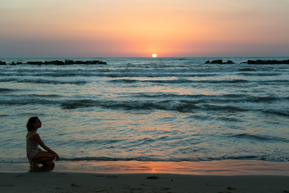 Person in blauen Jeans sitzt bei Sonnenuntergang am Strandufer