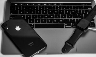 A set of electronic devices, including a black smartphone with a visible logo, a laptop with a sleek design and backlit keyboard, and a smartwatch with a dark screen, are arranged closely together.