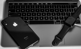 A set of electronic devices, including a black smartphone with a visible logo, a laptop with a sleek design and backlit keyboard, and a smartwatch with a dark screen, are arranged closely together.