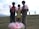 Three young boys standing on a grassy field, each holding pink cotton candy. There is a basket full of more cotton candies on the ground in front of them. The background shows a cloudy sky with distant mountains and a couple of people walking.