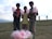 Three young boys standing on a grassy field, each holding pink cotton candy. There is a basket full of more cotton candies on the ground in front of them. The background shows a cloudy sky with distant mountains and a couple of people walking.