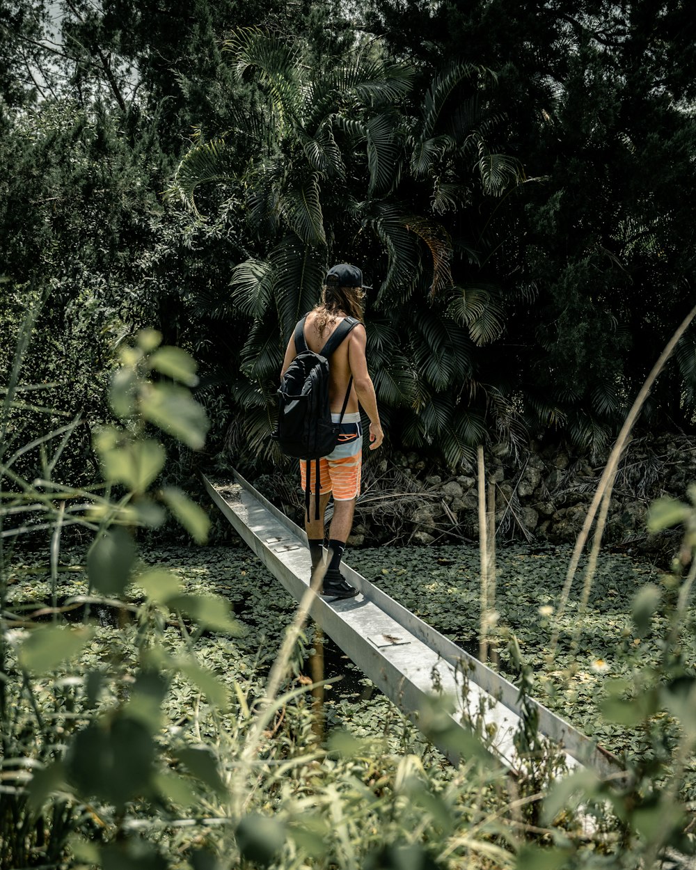 woman in orange and black backpack standing on brown wooden bridge surrounded by green plants during