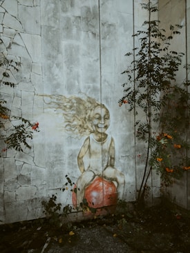 A mural painted on a textured stone wall features a joyful child with flowing hair rendered in a whimsical style, sitting atop a large red ball. Surrounding the mural are green plants and vines with clusters of small red flowers, adding a natural frame to the artwork.