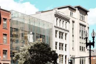 A contemporary glass-fronted building is visible, which features a large, prominent logo of a technology company. This structure is adjacent to older, more classical architecture, showcasing a contrast between modern and traditional styles. Trees and street lamps occupy the foreground, adding to the urban setting. The sky is clear with only a few clouds.