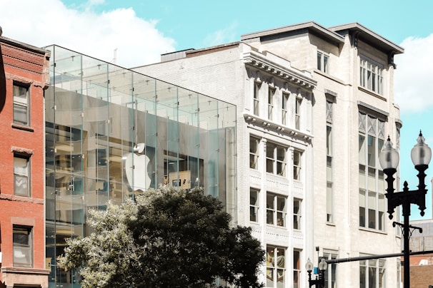 A contemporary glass-fronted building is visible, which features a large, prominent logo of a technology company. This structure is adjacent to older, more classical architecture, showcasing a contrast between modern and traditional styles. Trees and street lamps occupy the foreground, adding to the urban setting. The sky is clear with only a few clouds.