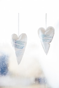 Two heart-shaped paper decorations with printed text hang from strings against a light, blurred background. The paper appears slightly translucent, and the hearts are gently curved with visible stitching along the edges.