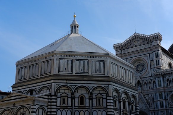 A detailed view of architectural buildings featuring intricate designs and patterns. The structure in the foreground has a distinct octagonal shape with a pointed dome, adorned with symmetrical motifs and a harmonious color palette. In the background, a larger building showcases a circular window with decorative elements and a cross on top.