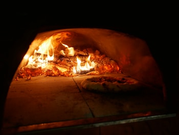A wood-fired oven with a roaring flame and burning logs creating a vibrant and warm atmosphere. A pizza with toppings is being baked on the oven floor beside the fire.