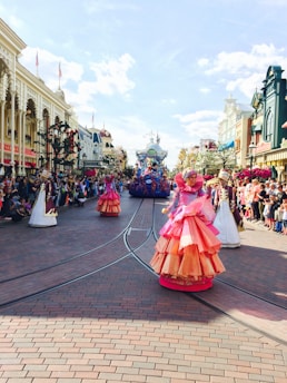 A vibrant parade with performers in elaborate, colorful costumes marching down a broad, brick-paved street lined with spectators. The parade features performers dressed in pink and orange outfits, and there are floats and decorations visible in the background. The crowd stands on either side of the street, with historic buildings adorned with flags and flowers.