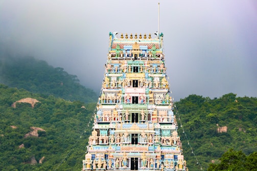 A richly decorated, colorful temple gopuram rises against a backdrop of lush green hills and a misty sky. Intricately carved sculptures and bright colors adorn the tower's multiple tiers.
