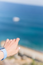 A person's hand is reaching out towards the sea, wearing a light blue smartwatch. The background features a blurred ocean with varying shades of blue, indicating a distant horizon.