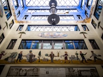 A colorful atrium with a large skylight, featuring a mural on the wall depicting various figures engaged in everyday activities. Palm trees and abstract designs add to the artistic feel. Architectural features include numerous windows and a prominent chandelier.
