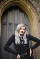 A person with long, light hair wearing a black top stands confidently in front of an arched wooden door set in a stone wall. The composition includes Gothic architectural elements.