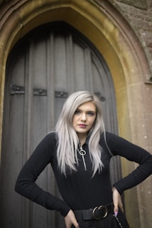 A person with long, light hair wearing a black top stands confidently in front of an arched wooden door set in a stone wall. The composition includes Gothic architectural elements.